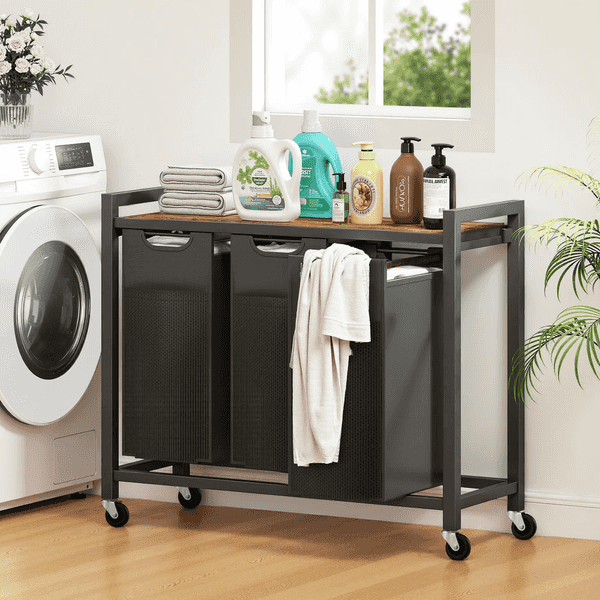 A black metal rolling laundry cart with a wood-look top shelf, shown in a bright laundry room next to a washing machine. The cart holds three black hamper bins, with a folded white towel and several laundry detergents and soaps arranged on the top shelf.