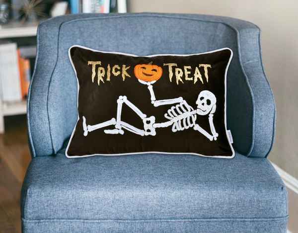 A rectangular black throw pillow with a white piped border sits on a blue upholstered armchair. The pillow features a white skeleton illustration reclining with an orange jack-o&