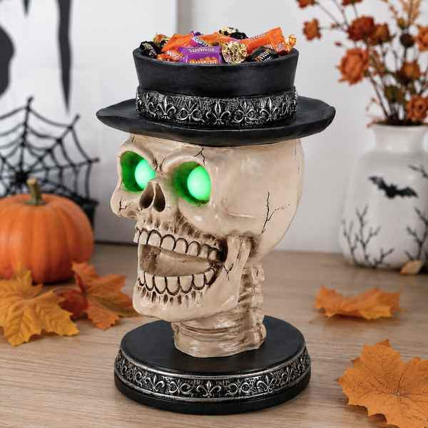 Halloween skull candle holder with glowing green eyes, black top hat with silver fleur-de-lis design, filled with Halloween candies, autumn leaves, pumpkin, and spider web decorations