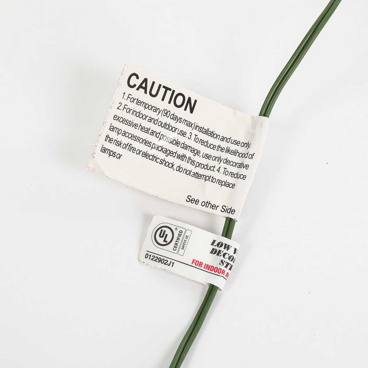 A close-up of a green electrical wire with two tags attached on a plain white background. The top tag is a white caution label with black text, and the bottom tag is a white UL certification label with red and black text.