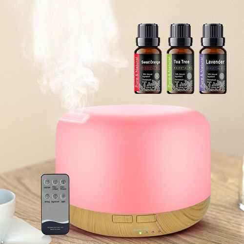 A pink and wood diffuser sits on a tabletop with steam rising from it, alongside a remote control. Three essential oil bottles labeled Sweet Orange, Tea Tree, and Lavender are shown above the diffuser, with a soft beige background.