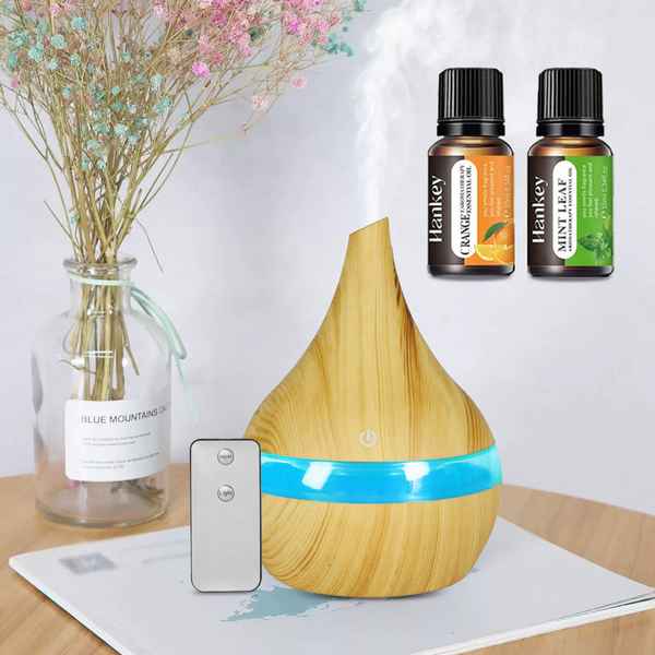 A teardrop-shaped wooden diffuser with a glowing blue band and visible mist, shown on a tabletop beside a remote control. Two small Hankey essential oil bottles are floating in the upper right, and a glass vase with dried flowers sits on the left.