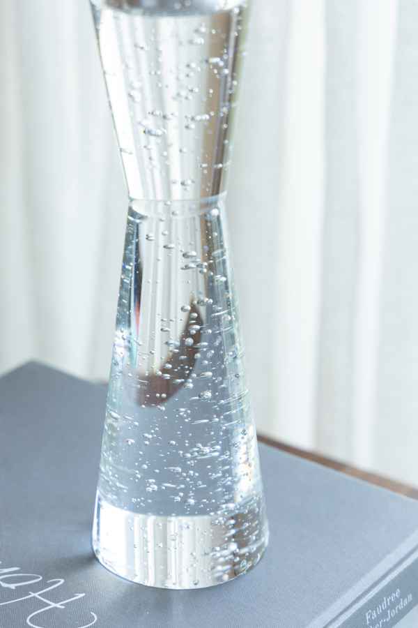 A clear glass hourglass-shaped vase filled with water and small bubbles, resting on a gray surface with a soft white curtain background.