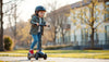 Young child wearing helmet rides three-wheel scooter in park with modern buildings