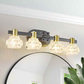 A modern four-light bathroom vanity light fixture with clear faceted glass shades, gold-toned caps, and a matte dark gray metal bar, mounted above a round mirror in a bright room.