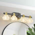 A modern four-light bathroom vanity light fixture with clear faceted glass shades, gold-toned caps, and a matte dark gray metal bar, mounted above a round mirror in a bright room.