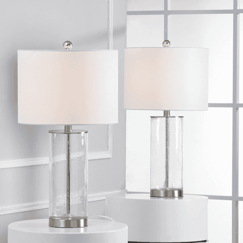 Two table lamps with white drum shades and clear cylindrical glass bases with silver metal accents, displayed on white pedestals in a bright interior setting.