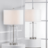 Two table lamps with white drum shades and clear cylindrical glass bases with silver metal accents, displayed on white pedestals in a bright interior setting.