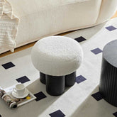 Round white ottoman on black legs in a living room setting with a coffee cup and saucer.
