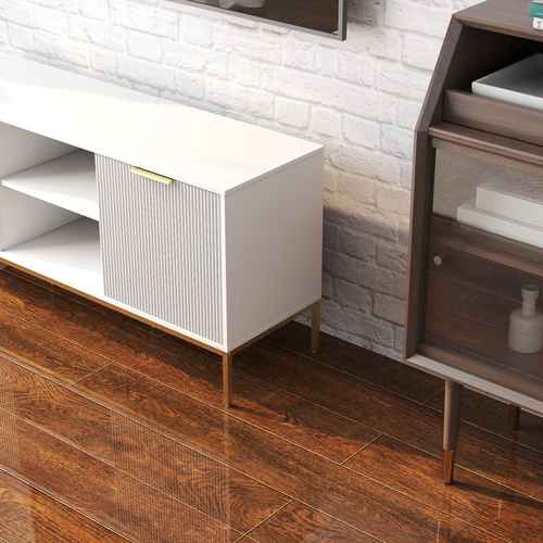 White TV stand with vertical ribbed doors and gold accents, on a wooden floor with a brown side table and a potted plant
