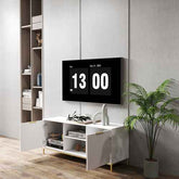 Modern white TV stand with vertical ribbed doors, gold accents, and gold metal legs, placed on a wooden floor with a potted plant nearby