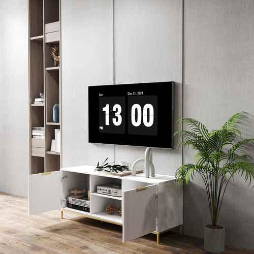 Modern white TV stand with vertical ribbed doors, gold accents, and gold metal legs, placed on a wooden floor with a potted plant nearby