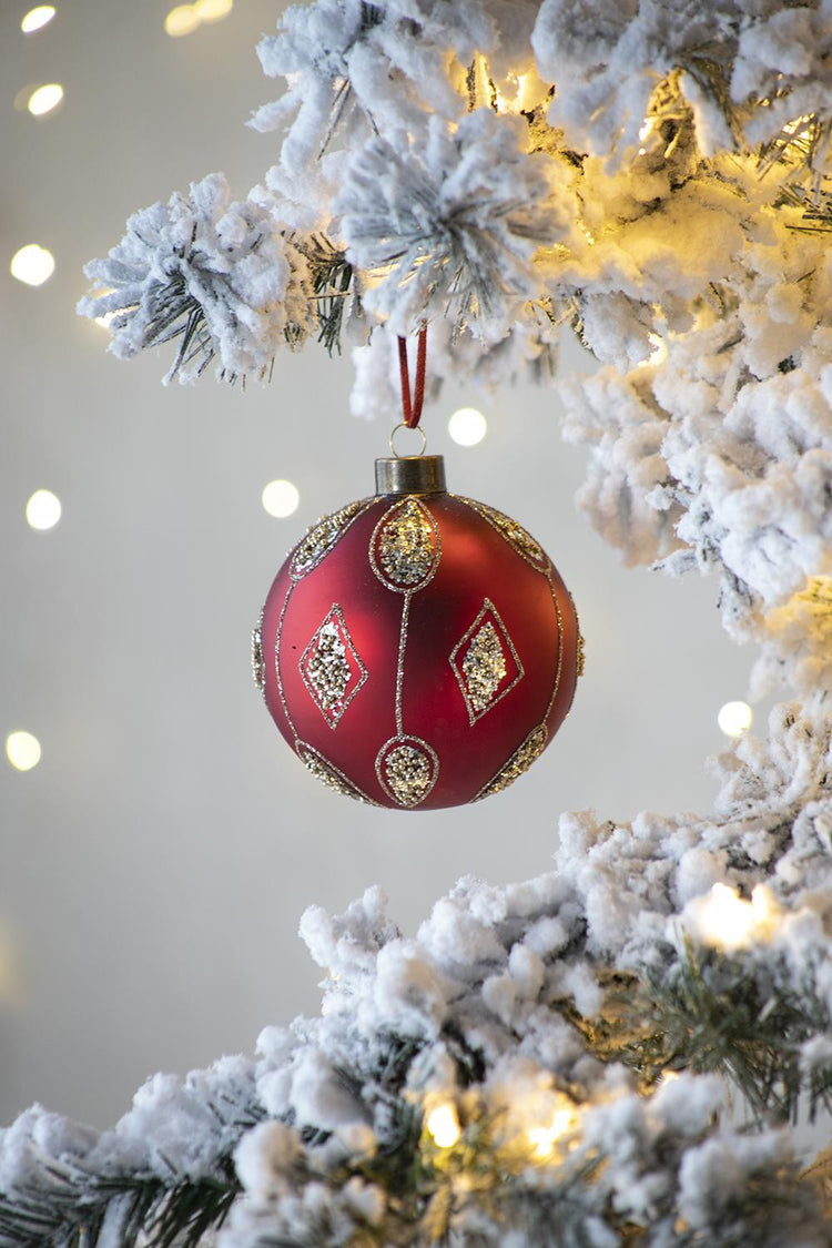 A red Christmas ornament with gold glittery diamond patterns hangs from a snowy flocked Christmas tree branch with warm white string lights in the background.