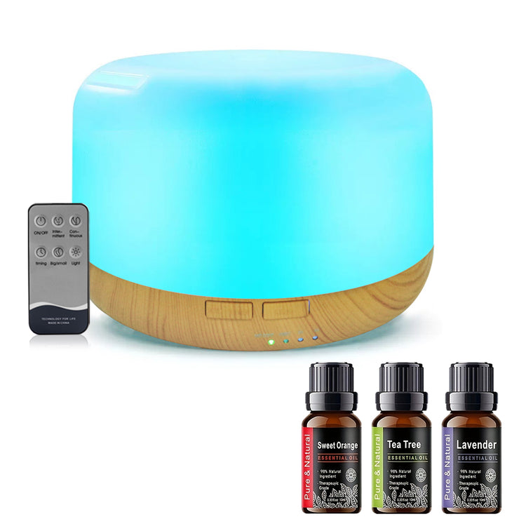 A cylindrical LED ultrasonic diffuser with a glowing blue light and a natural wood base, shown with a black remote control and three essential oil bottles labeled Sweet Orange, Tea Tree, and Lavender on a white background.