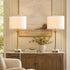 Two matching table lamps with white drum shades and gold bases are placed on either side of a console table in a styled interior room with a painting and vase.