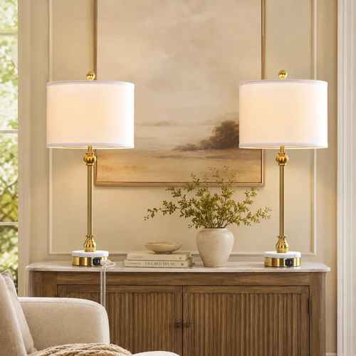 Two matching table lamps with white drum shades and gold bases are placed on either side of a console table in a styled interior room with a painting and vase.
