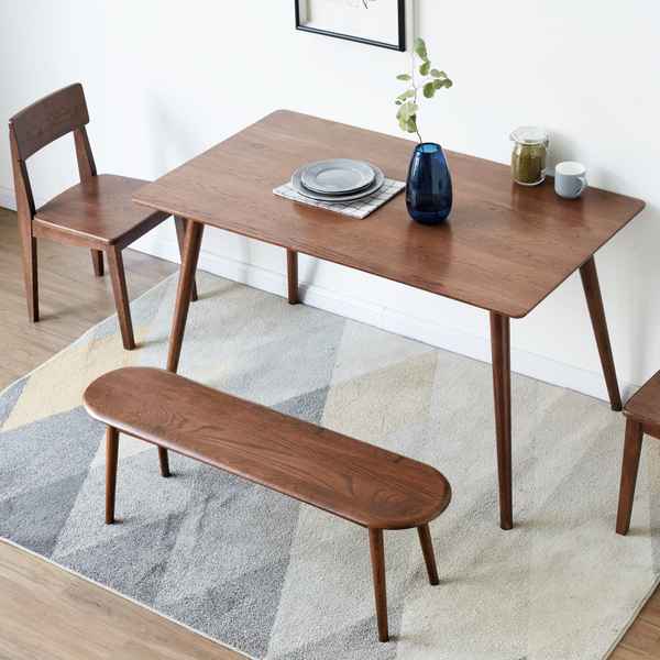 A mid-century modern wooden dining set featuring a rectangular table, a matching chair, and a long oval bench, all in a warm medium brown finish. The set is styled on a geometric patterned rug with a white wall and light wood floor in the background.