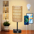 Wicker table lamp on a wooden surface with a plant, smartphone, and tablet in the background.