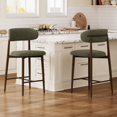 Two upholstered bar stools with dark wood legs and green-gray fabric seats and backs, shown in a bright kitchen with white cabinetry, marble countertops, and wood flooring.