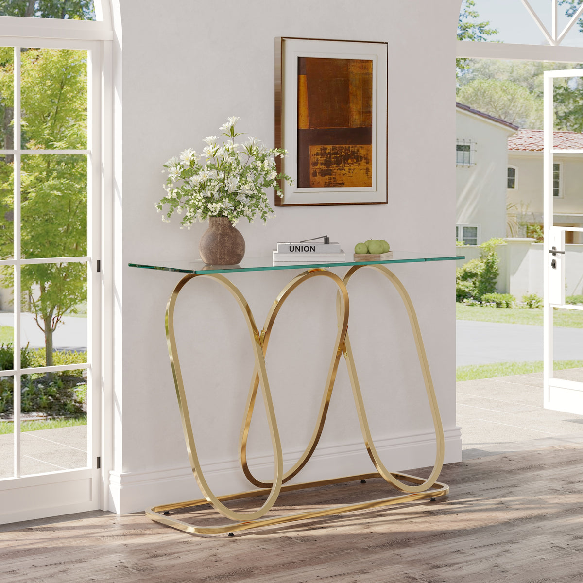 A modern console table with a clear glass top and a curved gold metal base, styled in a bright entryway with a vase of white flowers and framed art on top.
