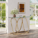 A modern console table with a clear glass top and a curved gold metal base, styled in a bright entryway with a vase of white flowers and framed art on top.