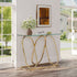 A modern console table with a clear glass top and a curved gold metal base, styled in a bright entryway with a vase of white flowers and framed art on top.
