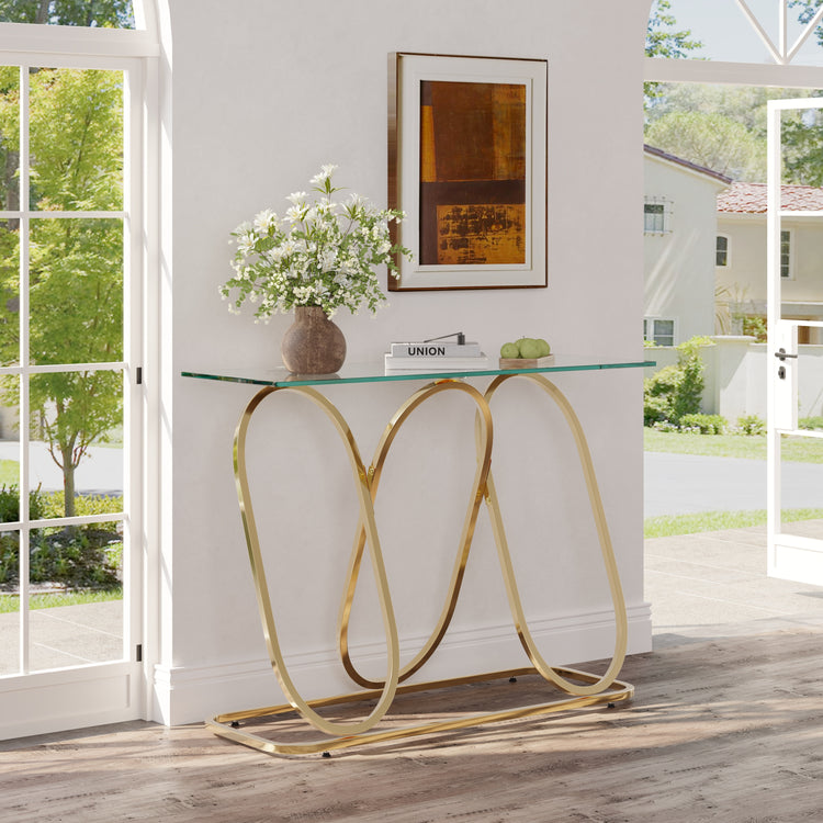 A modern console table with a clear glass top and a curved gold metal base, styled in a bright entryway with a vase of white flowers and framed art on top.