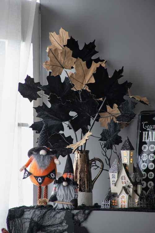 Halloween-themed tabletop decor arrangement featuring black and tan paper leaves, two small gnome figurines, a metal pitcher, a haunted house ornament, and a black advent calendar on a shelf against a gray wall.