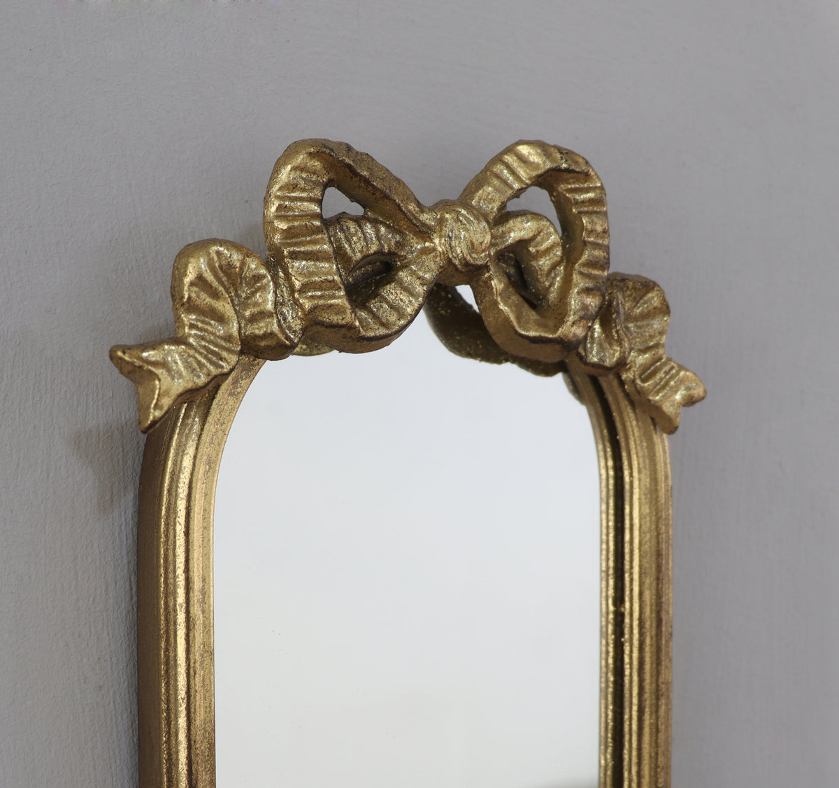 A close-up of an ornate gold mirror frame featuring a sculpted bow design across the top, set against a plain light gray wall.