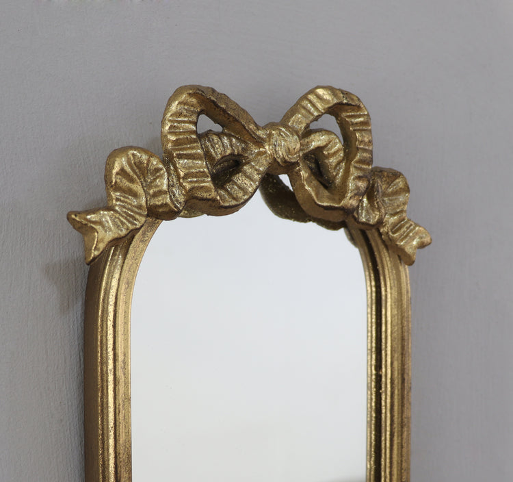 A close-up of an ornate gold mirror frame featuring a sculpted bow design across the top, set against a plain light gray wall.