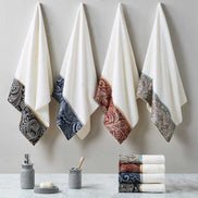 Four jacquard towel sets in black, navy, blue, and burgundy hanging on wall hooks, showcasing elegant cotton fabric and paisley borders.