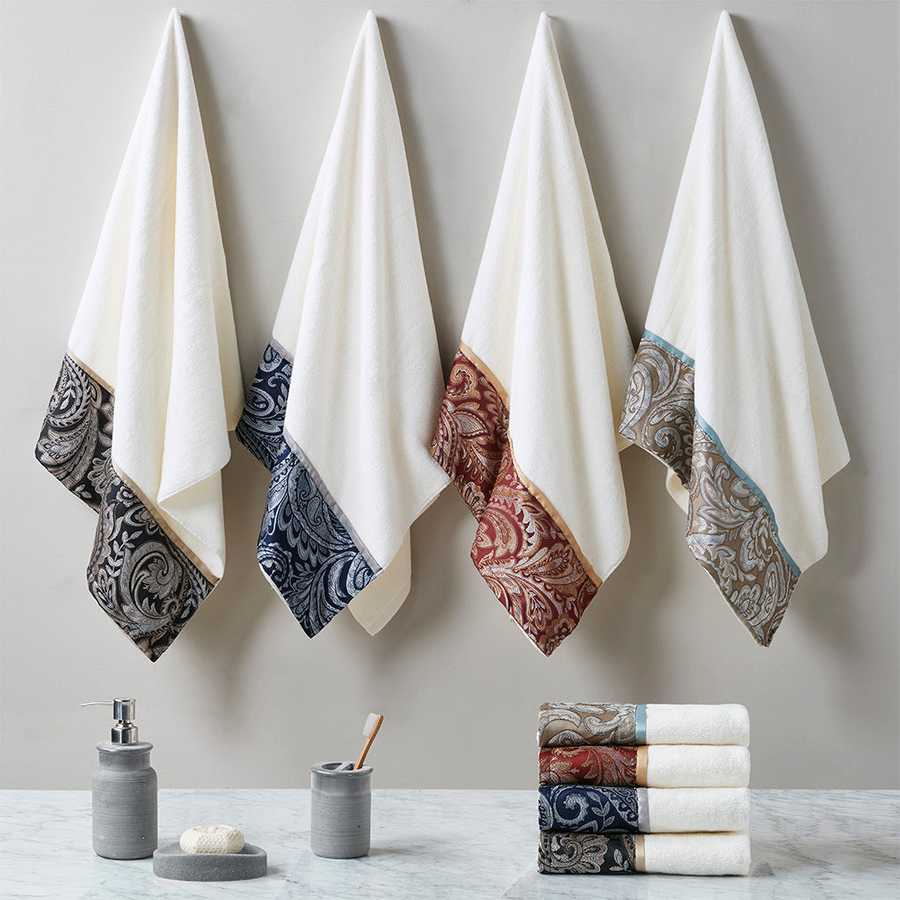 Four jacquard towel sets in black, navy, blue, and burgundy hanging on wall hooks, showcasing elegant cotton fabric and paisley borders.