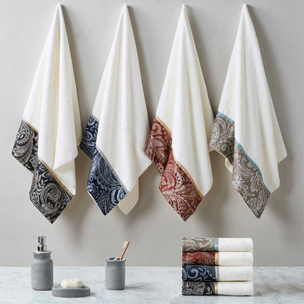 Four jacquard towel sets in black, navy, blue, and burgundy hanging on wall hooks, showcasing elegant cotton fabric and paisley borders.