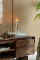 A lit candle in a clear textured glass holder sits on a dark wood sideboard, alongside a vintage camera and stacked books, against a warm beige wall.