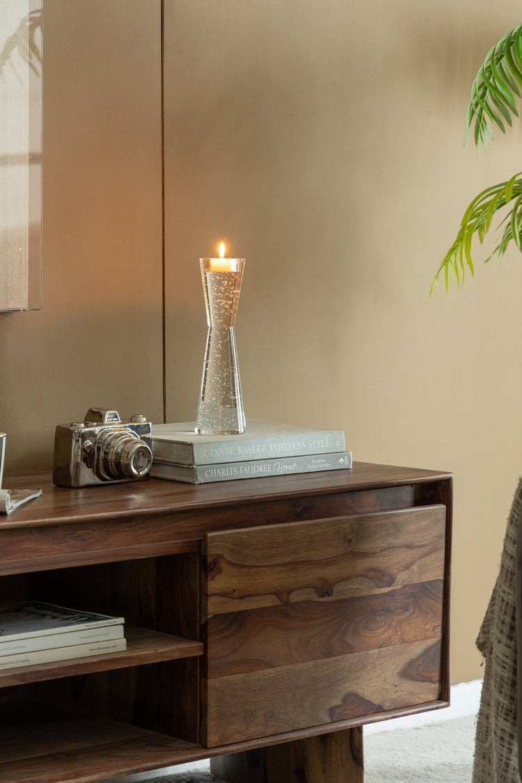 A lit candle in a clear textured glass holder sits on a dark wood sideboard, alongside a vintage camera and stacked books, against a warm beige wall.