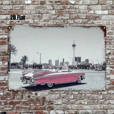 A rectangular metal wall art print mounted on a distressed brick wall. The print shows a vintage pink convertible car in front of a desert city skyline, with the sky rendered in grayscale and the car color highlighted in pink.