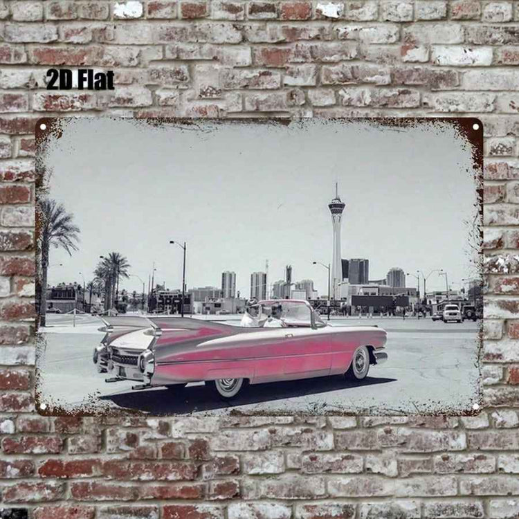 A rectangular metal wall art print mounted on a distressed brick wall. The print shows a vintage pink convertible car in front of a desert city skyline, with the sky rendered in grayscale and the car color highlighted in pink.