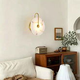 A modern wall-mounted sconce with a round white marble-look shade and curved gold metal arms, glowing warmly above a wooden sideboard in a styled living room.