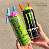 Two Monster Energy drink cans with colorful silicone straw covers, held in hands against a neutral concrete background.