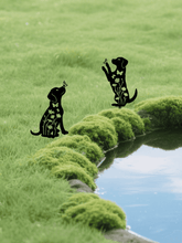 Two black dog-shaped garden markers with floral designs on a grassy area by a water body.