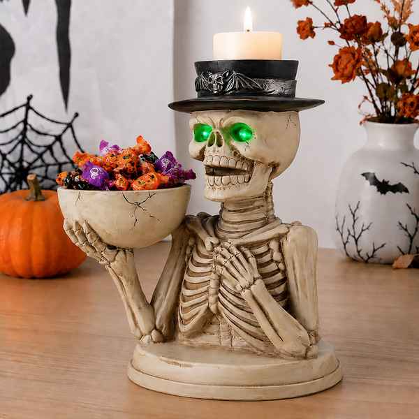 Halloween skeleton candle holder figurine holding a bowl of candy, with a lit white candle on its hat, displayed on a wooden table with a pumpkin and decorative vase in the background.
