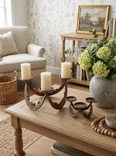A rustic three-light metal candelabra set displayed on a wooden coffee table in a cozy living room. The set includes a larger dark bronze candelabra with three white pillar candles and a smaller matching three-light holder.