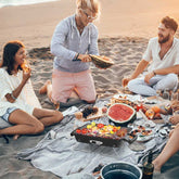 Group of people having a picnic on a sandy beach with food and drinks.