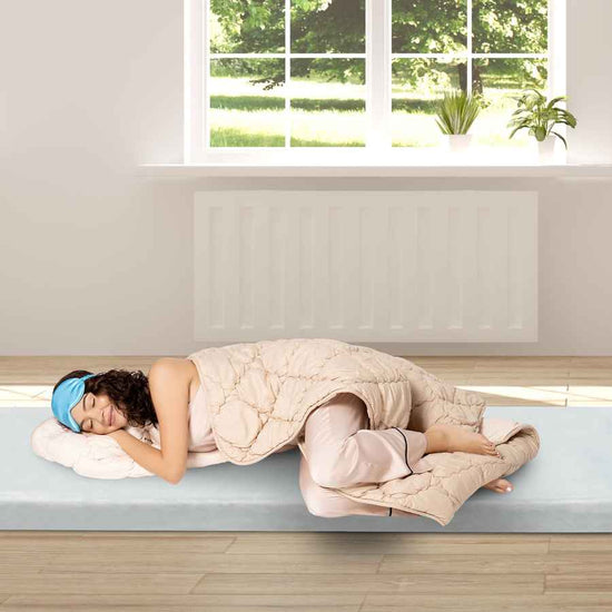 Person lying on a bed with a blanket and sleep mask in a bright room with a window.