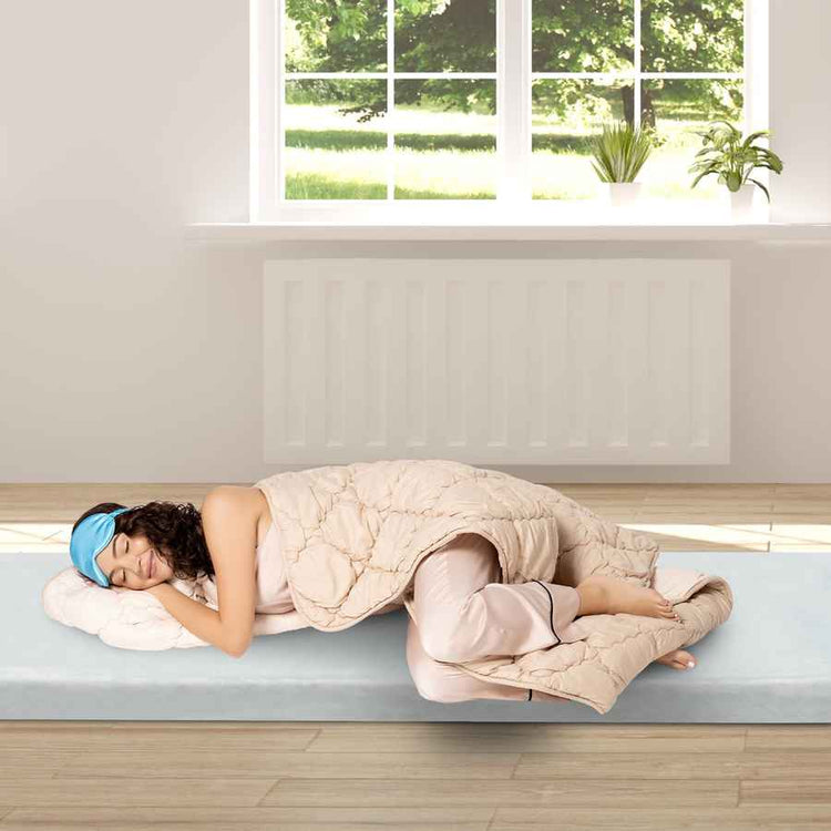 Person lying on a bed with a blanket and sleep mask in a bright room with a window.