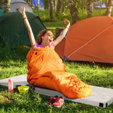 Person in an orange sleeping bag sitting on a camping mat with tents in the background