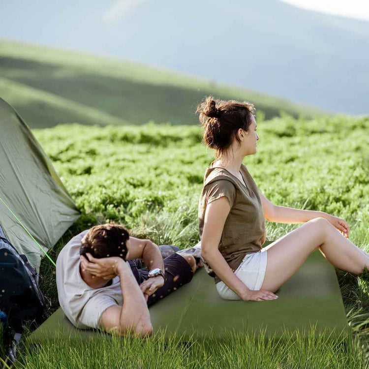 Two people sitting on a green camping mat in a grassy field with a tent and mountains in the background.