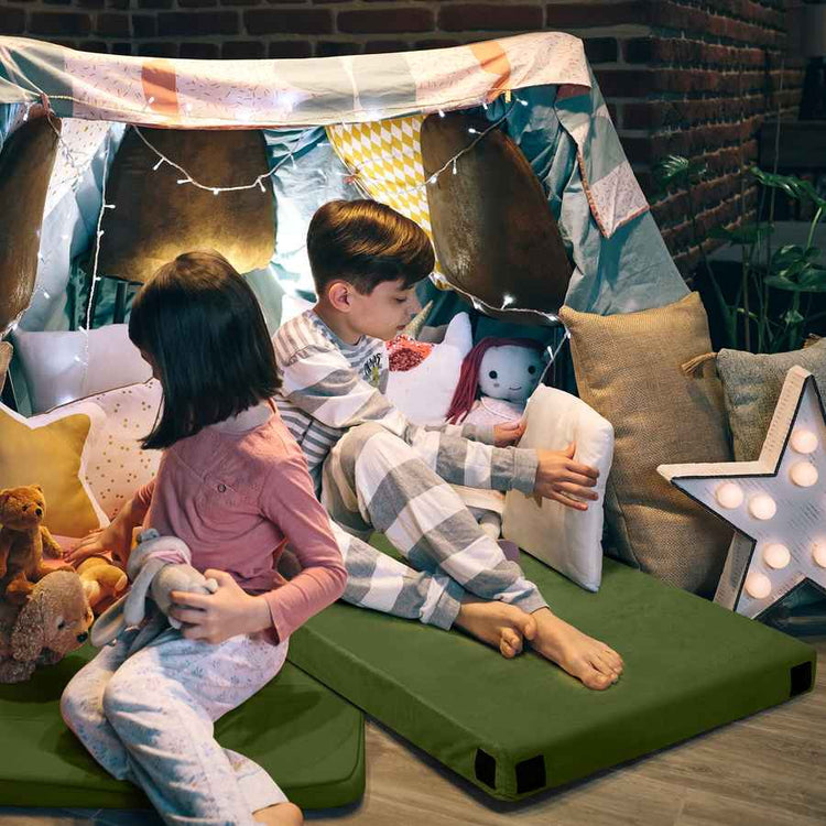 Two children playing with stuffed animals on a green foam play mat inside a makeshift fort made of blankets and pillows.