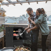 Man and woman grilling on a rooftop with cityscape in the background