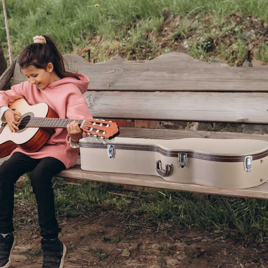 Child playing guitar next to an open guitar case on a wooden bench outdoors.